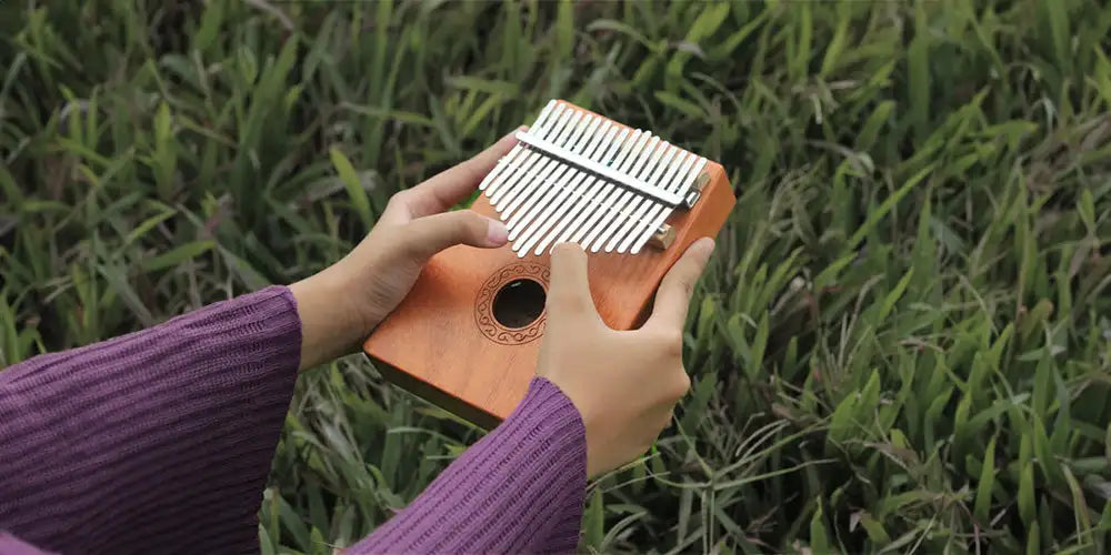 A wooden kalimba with silver-colored metal tines and a circular sound hole, held in hands wearing purple ribbed sleeves.